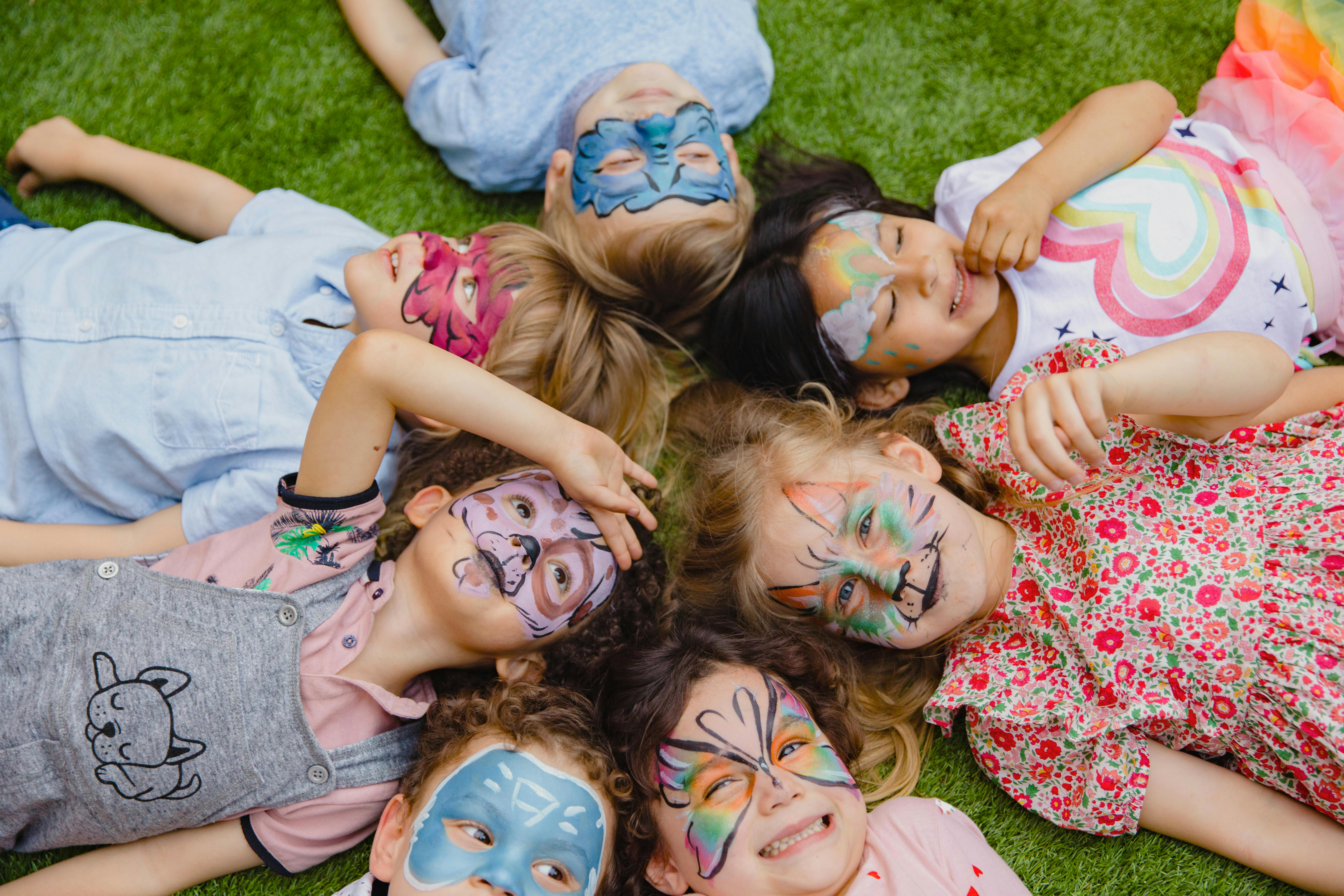 Children with colorful face paint lying together on grass, smiling and enjoying creative play